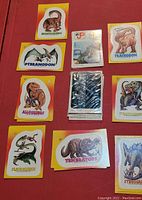Photo of various individual dinosaur stickers from the 1988 TOPPS Dinosaurs Attach Sticker Set laid out on a red surface, showing different dinosaur names and colorful cartoon artwork.