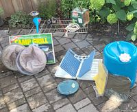 Full view of assorted camping gear laid out on stone patio including bug hats, ponchos, water jug, funnel, dish rack, cooking rack, and Coleman camp grill box.