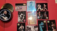 Photo showing 9 Elvis Presley trading cards displayed on a red surface, some cards in protective sleeves, alongside a round metal tin container for coasters.