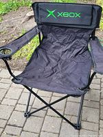 Folding camping chair with Xbox logo visible on backrest, showing black fabric, metal frame, and armrest cup holder