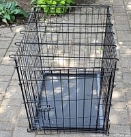 Front angled view of black metal dog cage with removable plastic tray on paved ground outdoors.