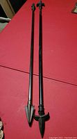 Two black metal extendable curtain rods lying on a red surface, each with a decorative spearhead or arrowhead shaped finial at one end.