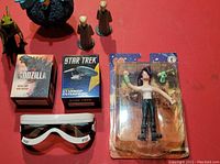Overview photo showing all sci-fi collectibles including boxed Godzilla set, boxed Star Trek starship model, Futurama Leela figure in packaging, white Star Wars Stormtrooper goggles, and several small Star Wars figurines on red surface.
