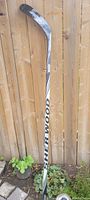 Full view of signed Sher-Wood hockey stick leaning against wooden fence outdoors with plants at base.