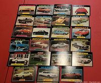27 vintage Topps car trading cards laid out on a red surface showing a variety of vintage car models including Brougham, Gremlin, TR7, Cordoba and others.