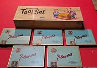 Five Player's Navy Cut Cigarette tin boxes arranged in a row with the Buildmaster wooden tool set box behind them, photographed on a red surface.