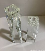 Front view of two clear crystal candle holders with cylindrical openings on top. Taller faceted prism shaped on left, shorter square shaped on right.