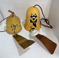 Two decorative bells with painted Native American inspired designs, one larger measuring about 9 inches, and one smaller with a dancer figure; both have hanging cords with windcatchers.