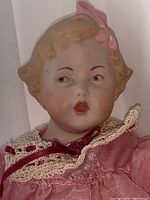 Front profile of bisque head doll with painted hair, pink bow, dressed in pink and cream lace with burgundy ribbon