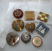 Overview of all nine vintage compact powder cases arranged on white fabric, showing various shapes and decoration styles.