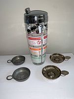 Photo of cocktail shaker with printed cocktail recipes placed between four wine tasting bowls, two silvered French cups and two pewter or similar metal cups.