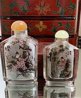 Pair of Chinese snuff bottles with detailed interior paintings, each with a different colored rounded stopper, set in front of lacquered box.
