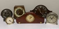 Collection of six clocks shown on flat surface including two small alarm clocks and four mantel clocks of various makes and wood and metal cases.