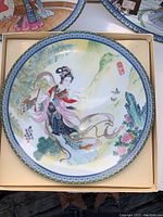 Top view of one decorative plate in its box showing Asian woman illustration with flowers and scenic background