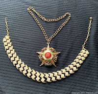 Two Coro necklaces on black textured background. One white enamel zigzag chain necklace 17 inches long. One gold-tone chain necklace 14 inches long with a large ornate star-shaped pendant featuring red and green enamel details.