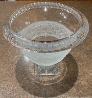 Clear crystal pedestal bowl with etched grape motif on the midsection, ribbed scalloped edge visible, set on a textured countertop.