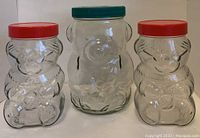 Three clear glass bear-shaped jars, one large with green lid and two smaller with red lids on a white background.