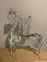 Front-left profile view of clear glass Pegasus figurine on wooden surface against white wall