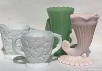 Four glass items on white surface: pink cornucopia vase, green jadeite vase, white creamer and sugar bowl, all milk glass variants.
