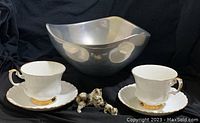 View of Nambe tri corner stainless steel bowl, two Royal Albert Val Dor teacups with saucers, and porcelain cat figurines.
