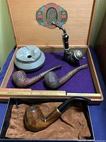 Photo of wooden cigarette box interior with purple fabric lining and multiple smoking pipes plus a pewter ashtray inside