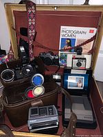 Panel inside suitcase showing multiple vintage cameras, Polaroid cameras including Swinger and Impulse Spectra, Ricoh F-34R, Canon AE-1 case, and photography book.