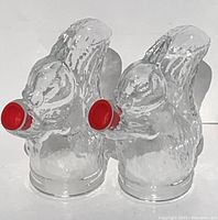 Pair of clear glass squirrel figurines from front side each holding red lid for dispensing candy, shown on white background.