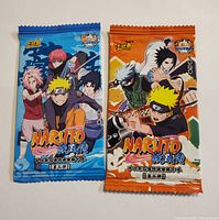 Front view of two sealed Naruto trading card packs, one blue and one orange, featuring characters from the anime.