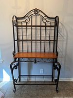 Full view of wrought iron and wood baker's rack showing overall design, black wrought iron frame with scroll details, wooden shelf in the middle and metal wire shelves above and below.