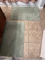 Two sage green bath mats laid out on tile flooring, showing overall size and condition.