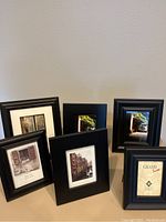 Six assorted black photo frames of various sizes arranged standing on a surface against a wall.