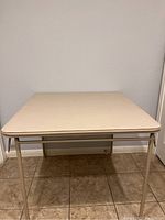 Front view of the card table showing the beige top and metal legs against a tiled floor and plain wall.