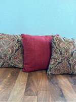 Photo of three decorative pillows arranged on wooden surface against a blue wall, two with ornate tapestry patterns and one solid red pillow in center.