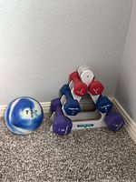 Image showing six neoprene-coated dumbbells in red (3 lb), blue (5 lb), and purple (8 lb) resting on a white Definity labeled stand, placed on carpeted floor against a wall corner.