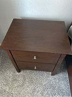 Top and side view of dark brown nightstand showing flat top, two drawers with metal knob handles, square legs on sides, placed on carpeted floor.