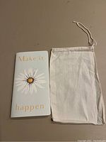 Closed image of the light blue Planner Perfect Journal with a daisy and gold text 'Make it happen' on the cover alongside a white muslin drawstring bag.