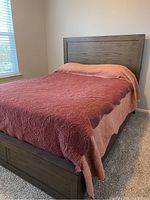 Full side view of queen bed with pink and red quilted bedspread, showing the wooden headboard against the wall and part of the footboard.