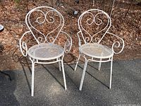 Two white painted wrought iron bistro chairs placed outdoors with fallen leaves in background, showing overall design and condition including visible rust.