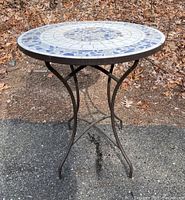 Full view of round bistro table with mosaic tile top and metal base outdoors on pavement with foliage background