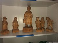 Six carved wooden figurines displayed on white shelving, including one larger figure and five smaller ones.