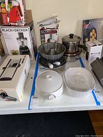 Photo showing a variety of kitchen items arranged on a table including boxed Black+Decker food chopper, Nutri Slicer box, stacked cake pans, Cuisinart pot, Sophie Conrad casserole dish, and CorningWare casserole dish.