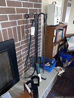 Fireplace tools set including poker, tongs, shovel on wrought iron stand in front of brick fireplace, with bookends, matches, and electronic chargers nearby