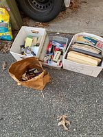 Photo of four containers on driveway including brown paper bag holding pens and pencils, plastic box with office supplies like tape dispensers and staplers, and two cardboard boxes with folders and paper items.