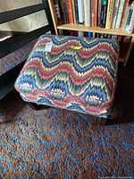 Ottoman with multicolor zigzag upholstery showing top and side view beneath bookshelf