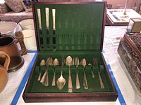 Opened wooden hinged case showing the green felt interior and arrangement of multiple silver plated flatware pieces including knives, forks, and spoons.