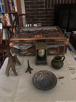 Photo showing the entire lot on a table, including a large German tin cookie tin with scenic designs, wooden horse statue, Asian decorative egg in case, small ceramic vase with initials, metal statue, ceramic dish, and small metal Eiffel Tower figurine.