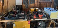 Wide view of assorted tools on a table including vice, jumper cables, clamps, staple tool, Big Jack, work lights, car jack, hammers, level, and metal ruler in a workshop setting.