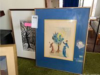 Two framed artworks, one colorful print with figures and tree accented by blue matting, and one black and white tree silhouette behind it.