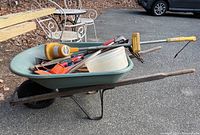 Green metal wheelbarrow filled with various tools including snow scrapers and a plastic tub with lid, placed outdoors on pavement
