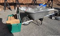 Side view of gray plastic True Temper wheelbarrow with foam-filled solid tire, green storage bin with wooden handles, metal mesh, and hardware on ground.